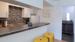 Una cocina moderna con salpicadero de piedra, armarios blancos y encimera gris. Hay dos taburetes amarillos y una planta pequeña en la encimera.