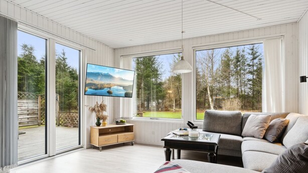 A spacious living room with a large sofa, a coffee table, and a TV mounted on the wall. There is a sliding glass door leading to an outdoor area with a wooden deck and a view of trees and mountains.
