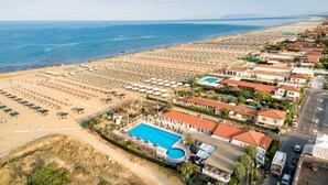 An aerial view of a coastal resort with a large swimming pool, beach umbrellas, and beach chairs.