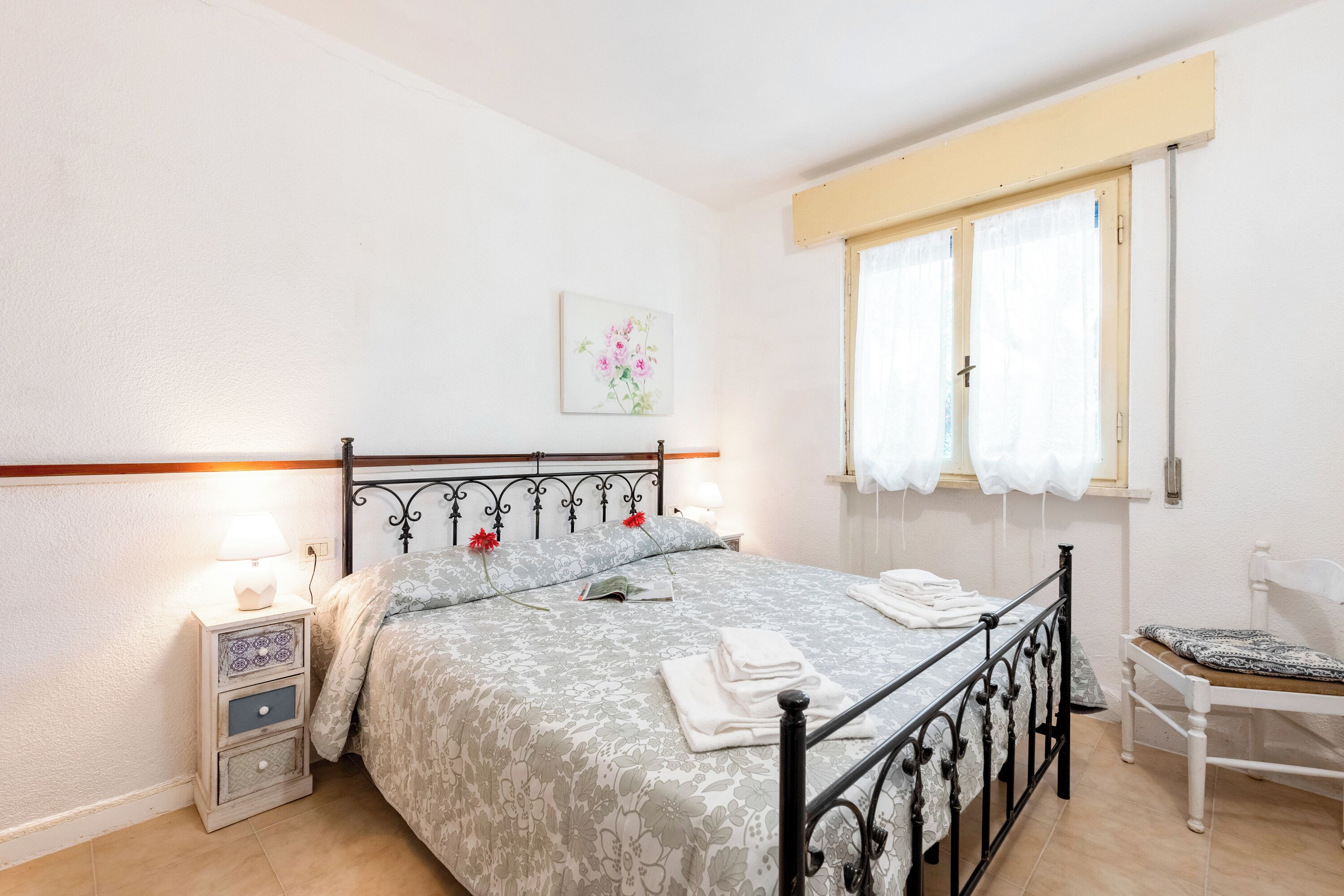 Un dormitorio con cama, mesita de noche, silla, ventana y una obra de arte en la pared.