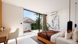 A modern bedroom with a large bed, a desk, and a view of a tropical garden through a sliding glass door.