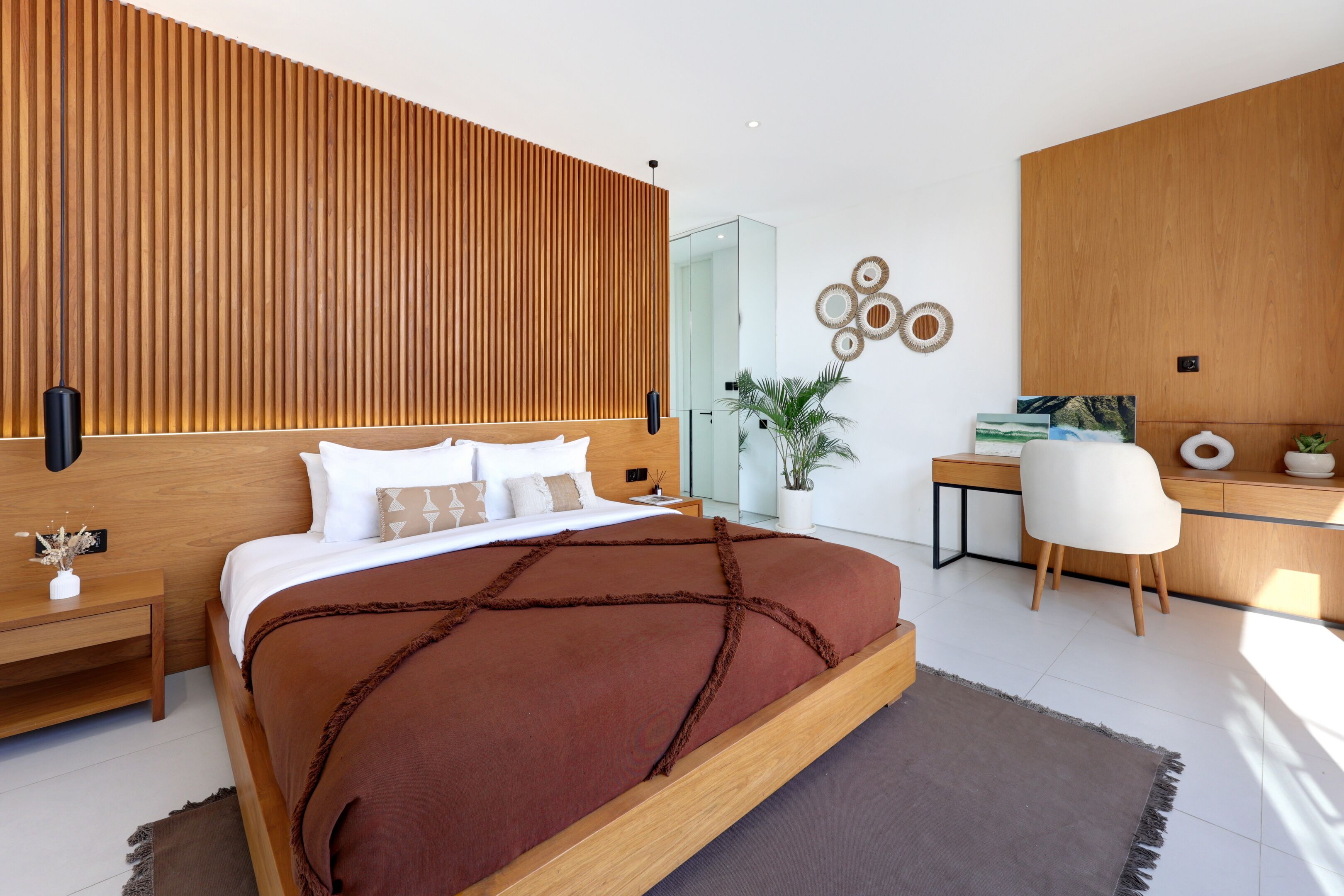 A modern bedroom with a wooden headboard, a large bed with a brown blanket, a desk with a chair, and a potted plant.