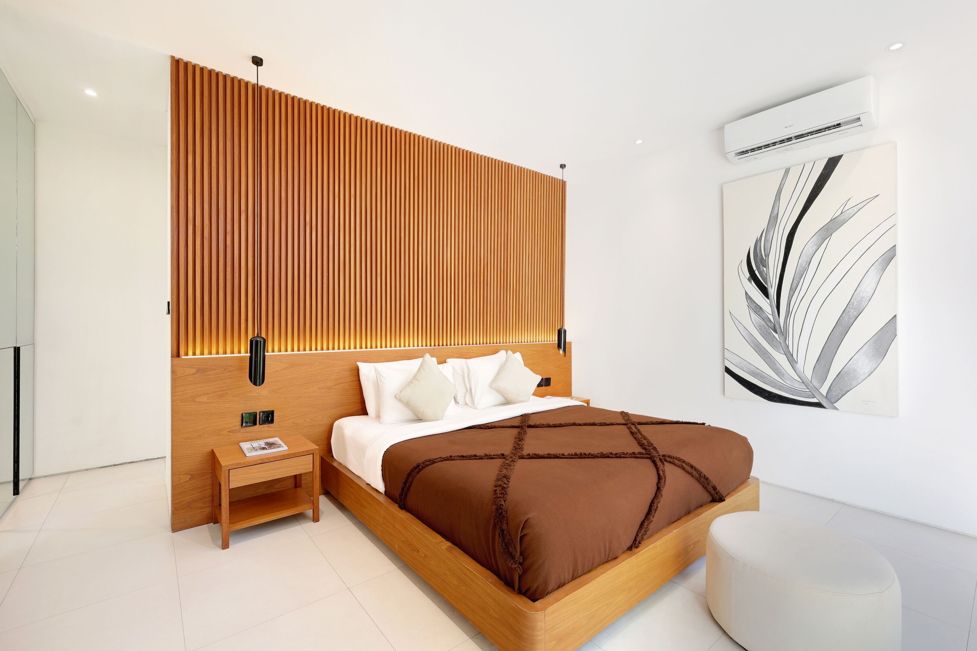 A modern bedroom with a wooden headboard, a bedside table, a large bed with a brown bedspread, a round ottoman, and a wall-mounted air conditioner.