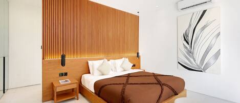A modern bedroom with a wooden headboard, a bedside table, a large bed with a brown bedspread, a round ottoman, and a wall-mounted air conditioner.
