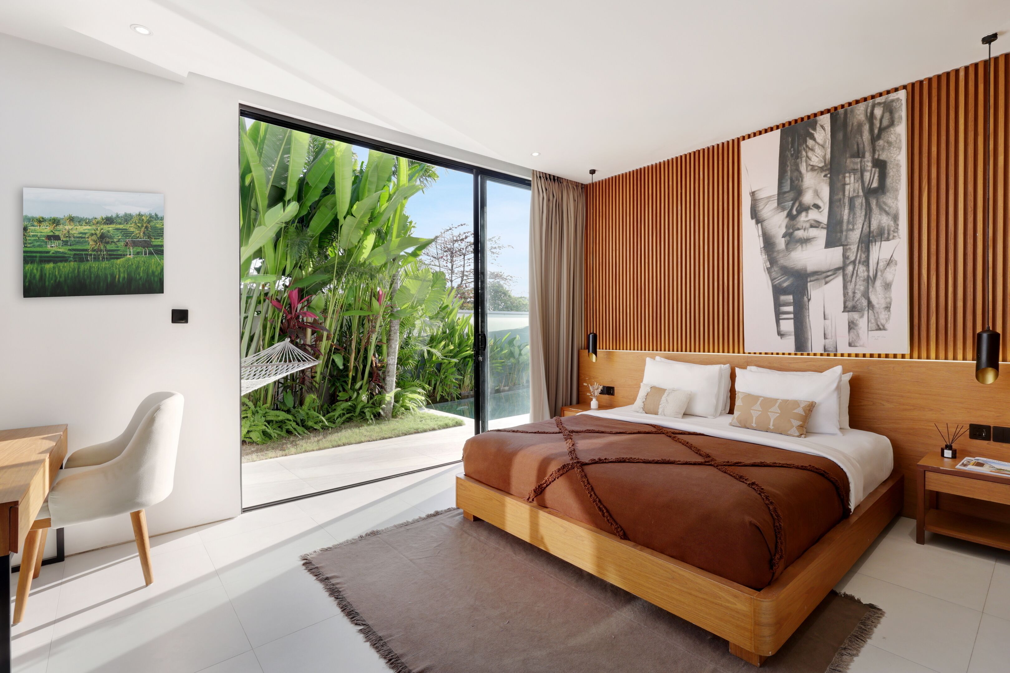 A modern bedroom with a large bed, a desk, and a view of a lush garden through a sliding door.