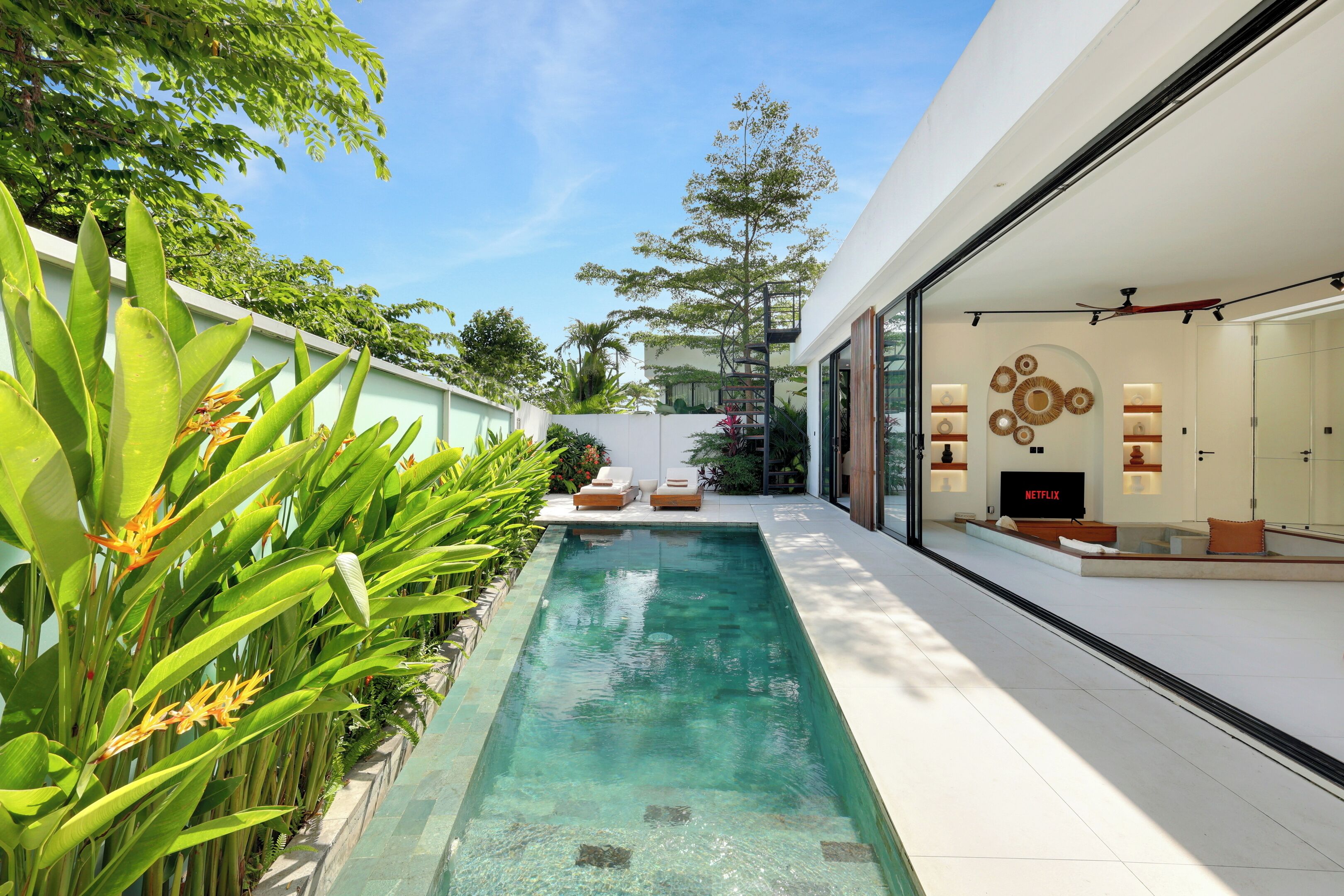A modern pool area with a clear pool, surrounded by lush greenery and a contemporary interior with a Netflix TV.