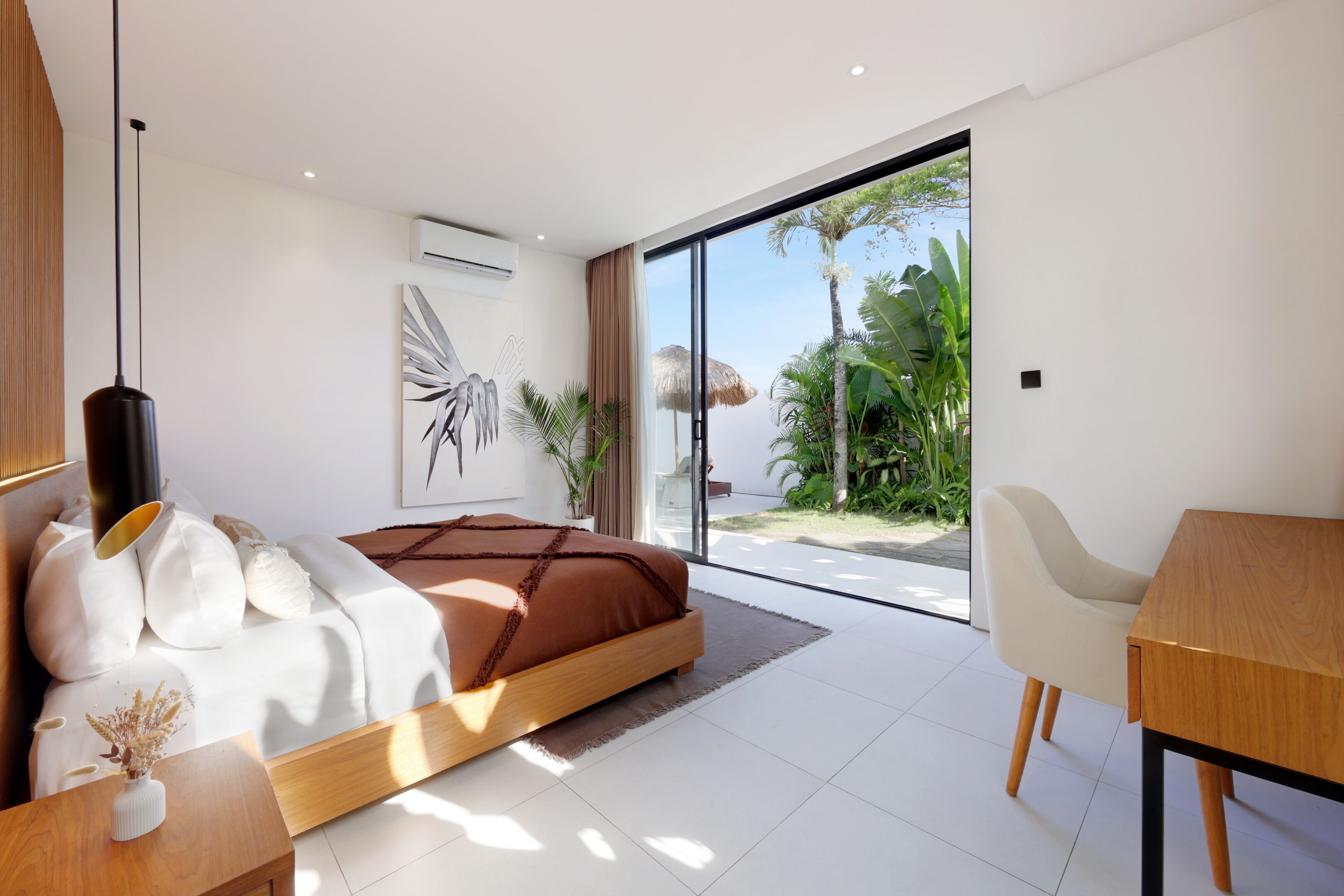 A modern bedroom with a large bed, a wooden bedside table, a white chair, and a view of a tropical garden through a glass door.