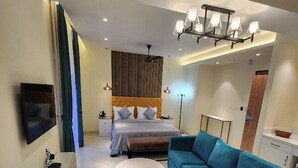 A modern hotel room with a bed, a blue sofa, a TV, a desk, and a chandelier. - Hotel Southgate Goa (Calangute)
