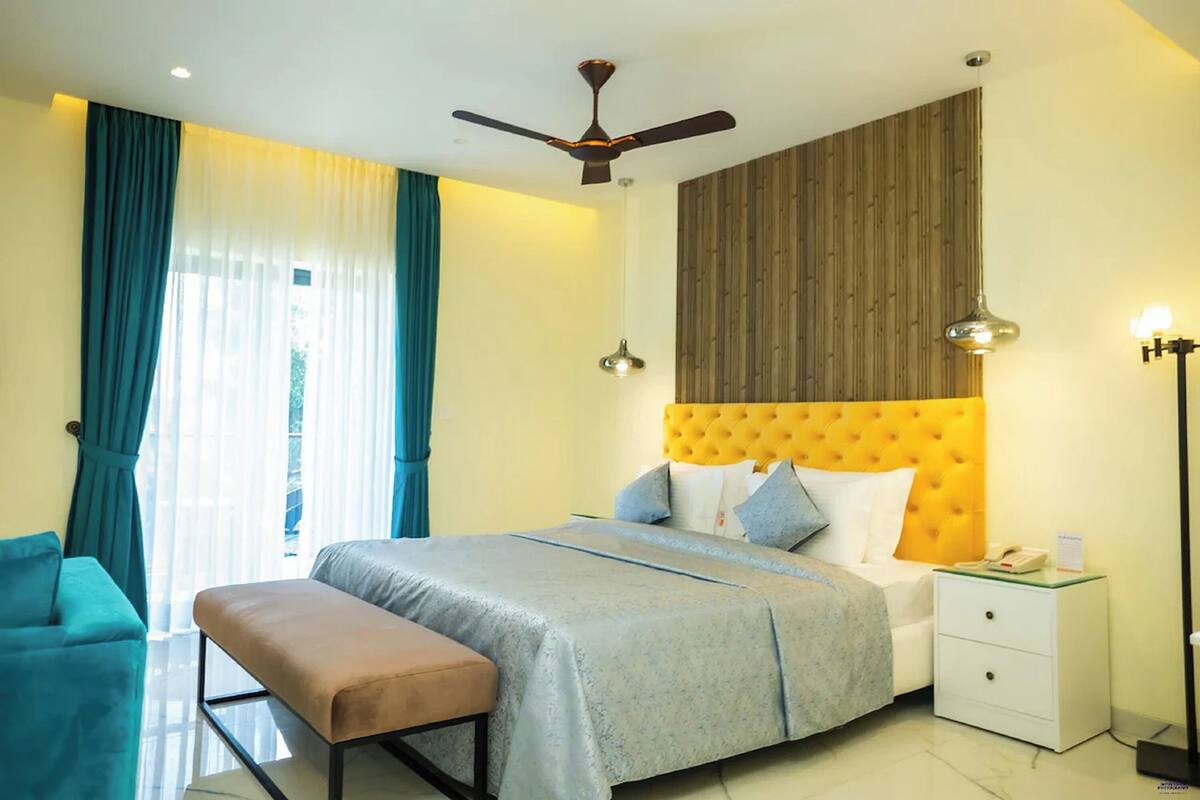 Habitación de hotel con cama, reposabancos, mesita de noche, ventilador de techo y ventana con cortinas.