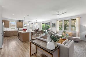 A modern kitchen with wooden cabinets, a dining area with a table and chairs, a living room with a sofa and a coffee table, and large windows with a view of the outdoors. - Pakele Oahu by AvantStay | 5 Min to Makaha Beach (Waianae)
