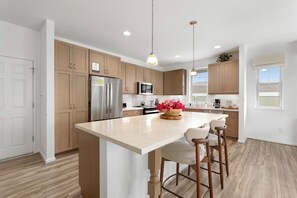 A modern kitchen with wooden cabinets, stainless steel appliances, a central island with bar stools, pendant lighting, and a view of the outdoors through a window. - Pakele Oahu by AvantStay | 5 Min to Makaha Beach (Waianae)