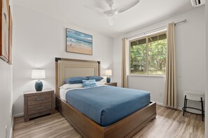 Une chambre à coucher comprenant un lit, des tables de chevet, un ventilateur de plafond, une fenêtre avec des rideaux et une décoration murale sur le thème de la plage.