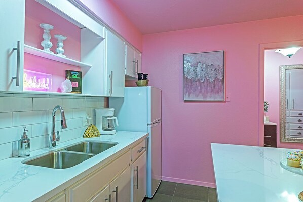 Una cocina con armarios blancos, un lavamanos y una encimera. Hay un refrigerador y una cafetera sobre la encimera.