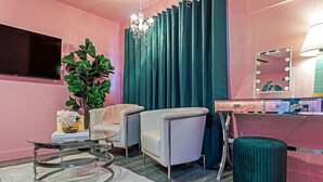 A pink-themed room with a TV, a plant, two chairs, a round table, a mirror with lights, and a green ottoman.
