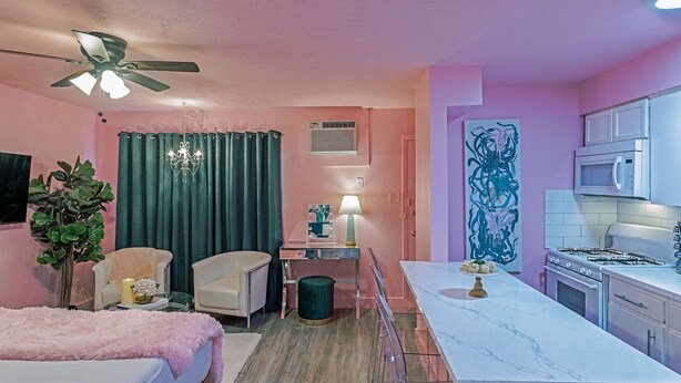 A compact room with a bed, a desk, a chair, a kitchenette, and a ceiling fan.
