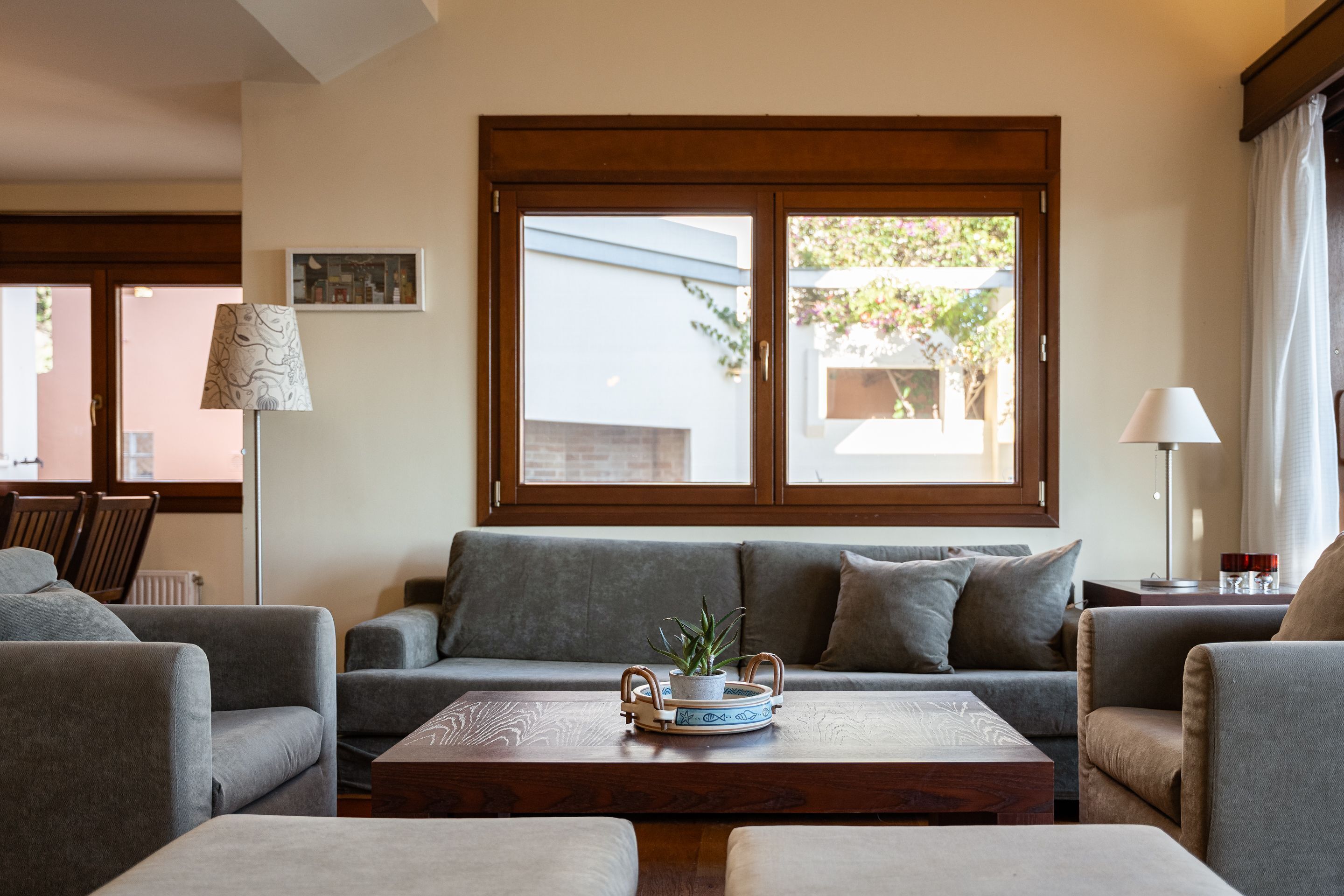 A living room with a grey sofa, a wooden coffee table with a plant, and a window with a view of greenery.