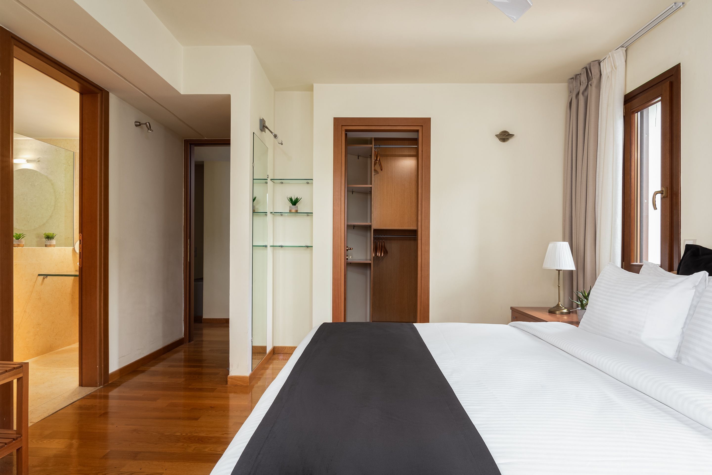 A hotel room with a large bed, a wooden wardrobe, a bedside table with a lamp, and a glass cabinet with plants.