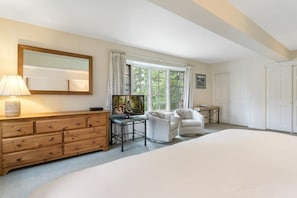 Um quarto com uma cama grande, uma cômoda de madeira, uma televisão sobre um móvel, duas poltronas brancas e uma janela com cortinas.