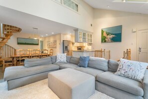 Amplia sala de estar con un sofá modular grande, un comedor con muebles de madera, una cocina con armarios blancos y una escalera que conduce a un nivel superior.