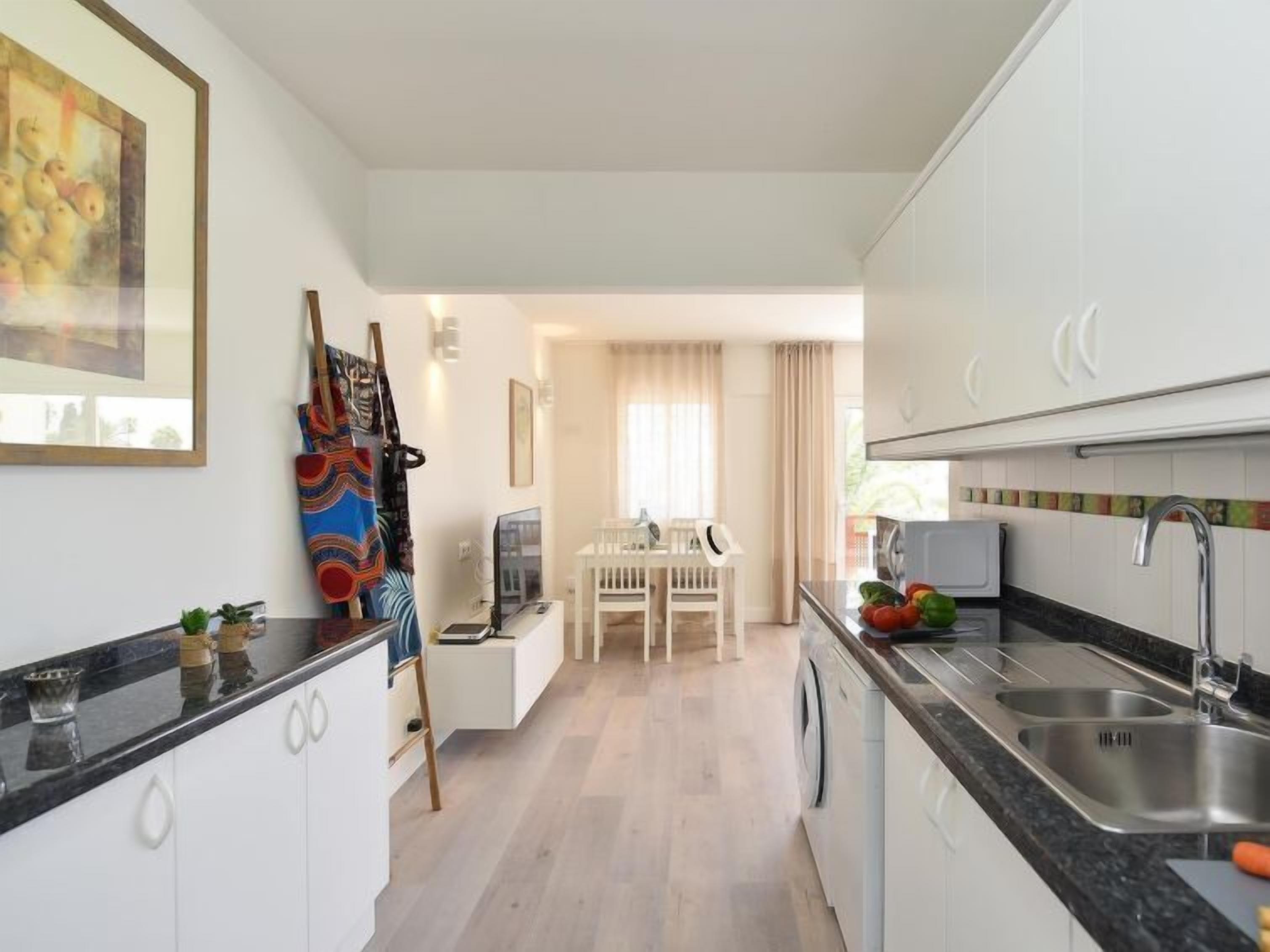 Una cocina moderna con armarios blancos, encimera negra y electrodomésticos de acero inoxidable. Hay una zona de comedor con una mesa y sillas blancas, y un cuadro en la pared.