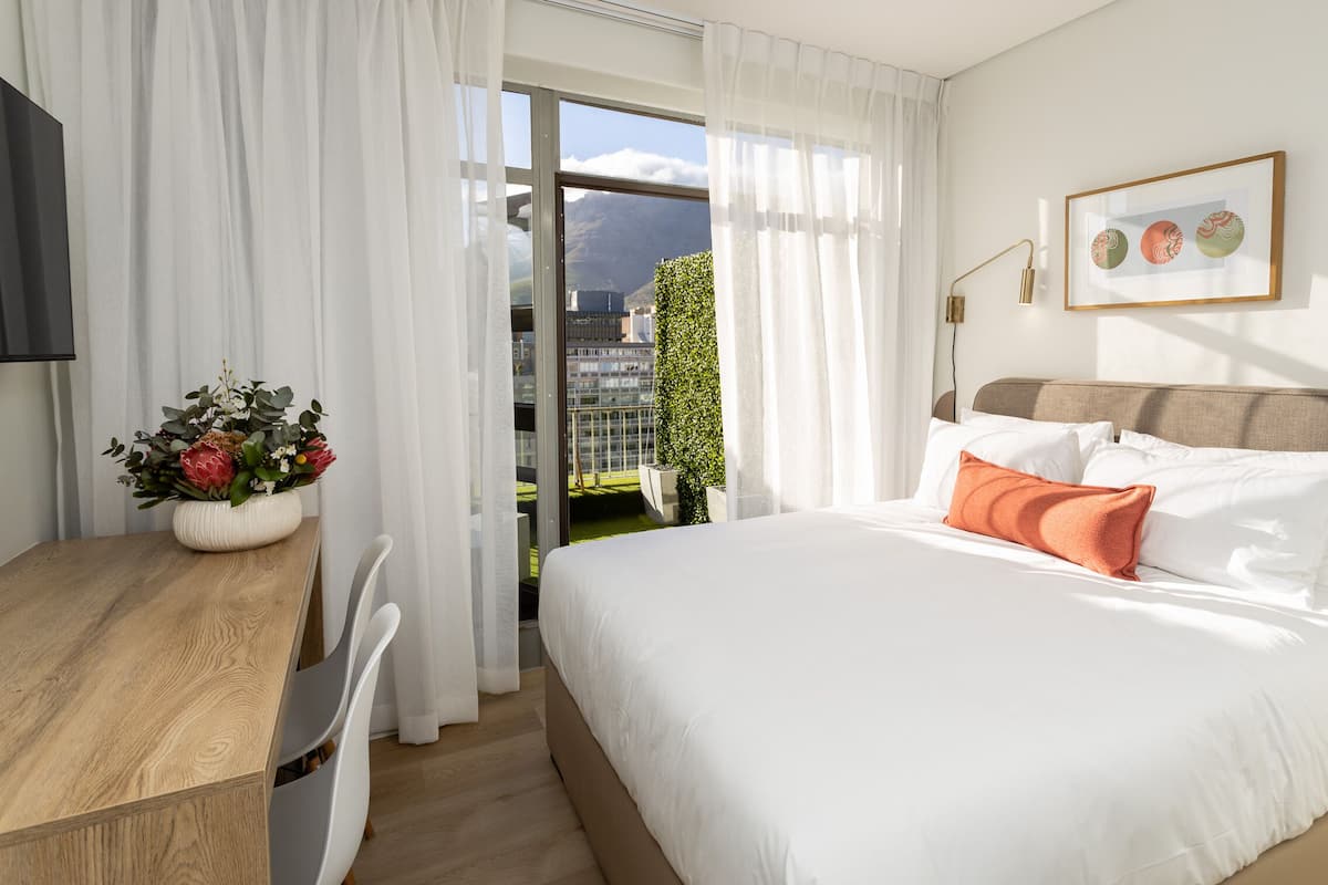 A hotel room with a bed, a desk, a chair, a window with curtains, and a view of greenery and buildings.