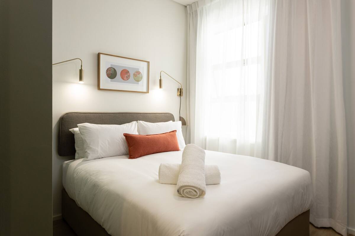 A neatly made bed with white linens, a brown headboard, and a single orange pillow. There are two wall-mounted lamps with gold accents, and a framed picture above the bed.