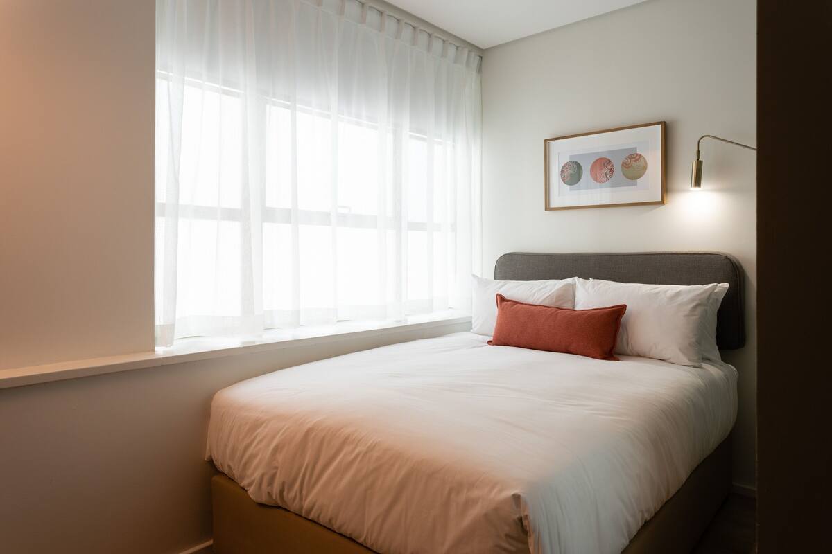 A neatly made bed with white and orange pillows, a framed picture on the wall, and a window with sheer curtains.