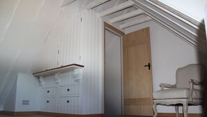 Una stanza con soffitto spiovente, una porta in legno, un armadietto bianco con cassetti e una sedia dai toni chiari.