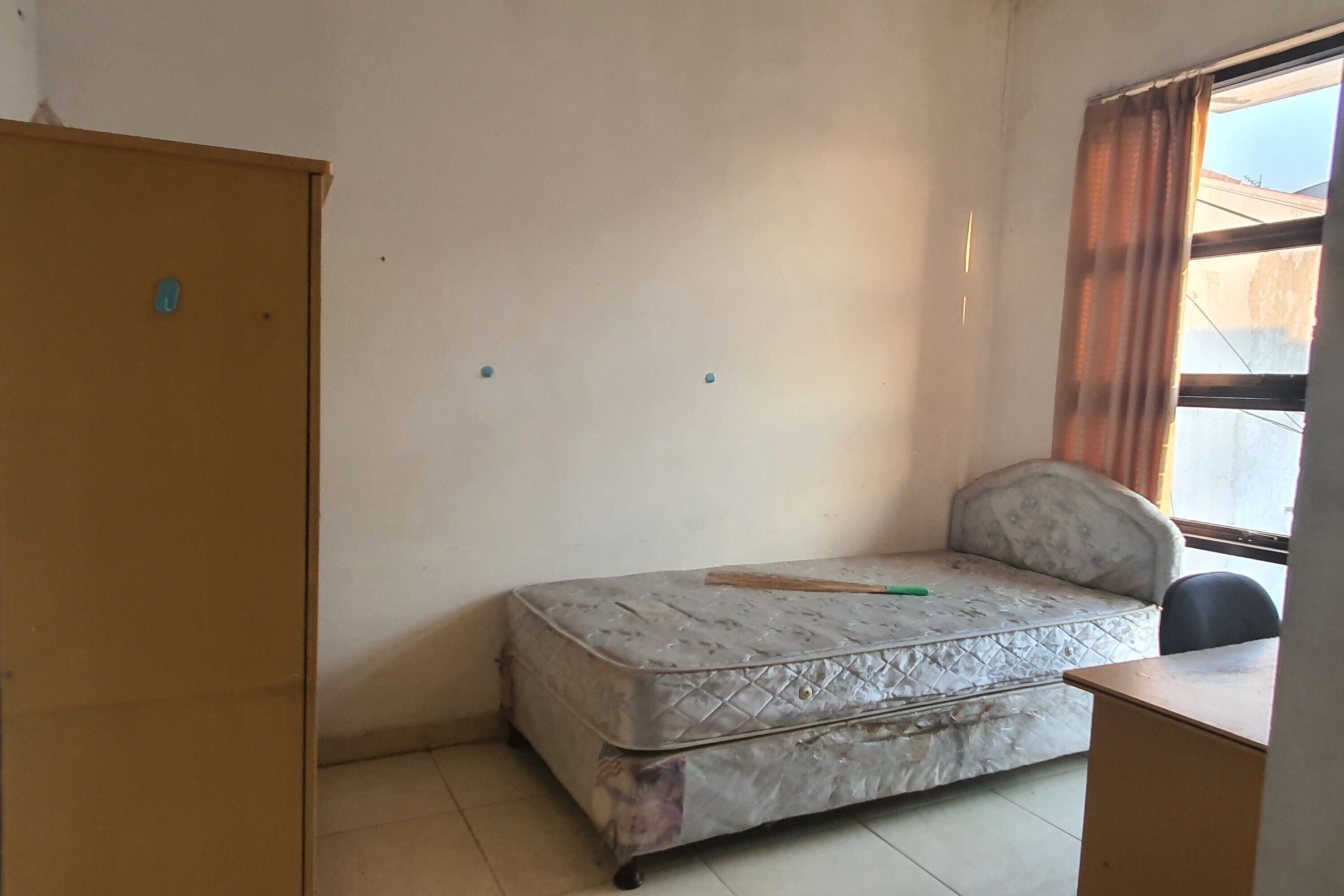 A small, sparsely furnished room with a single mattress bed, a wooden desk, and a wardrobe.