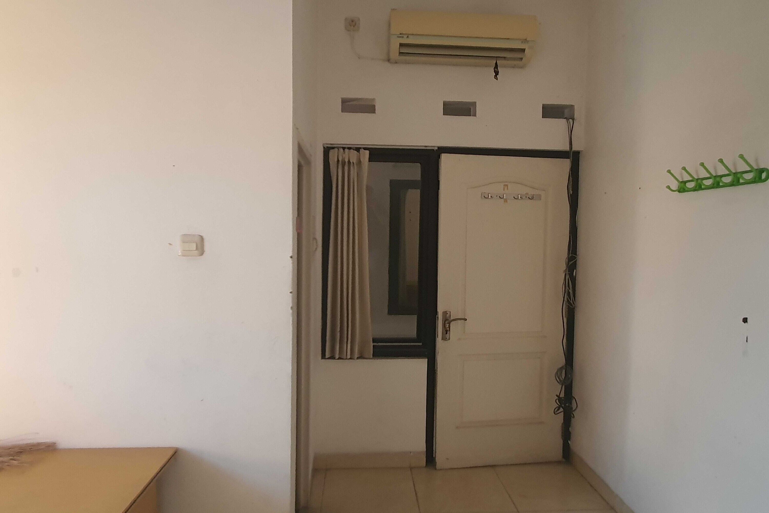 Quarto com porta branca, janela com cortinas, ar-condicionado e um cabideiro verde.