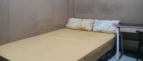 A small, single-bed room with a plain mattress and a wooden desk.
