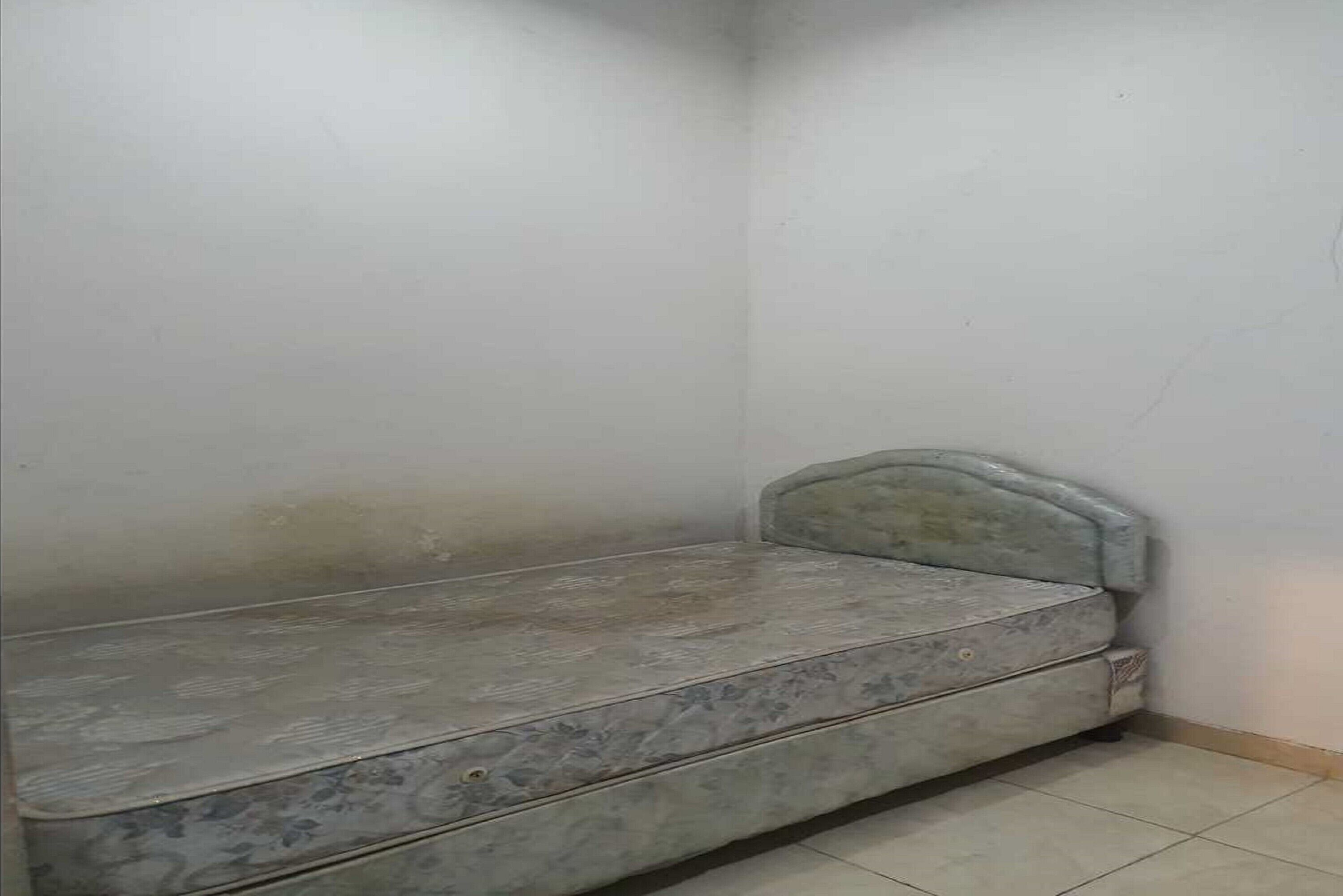 A small, single-bed room with a simple, unadorned mattress and headboard.