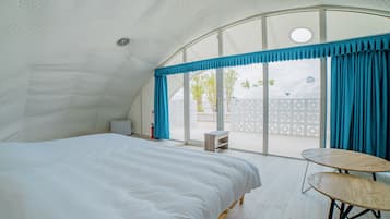 A bedroom with a large bed, a small table, and a view of the outside through a window with blue curtains.