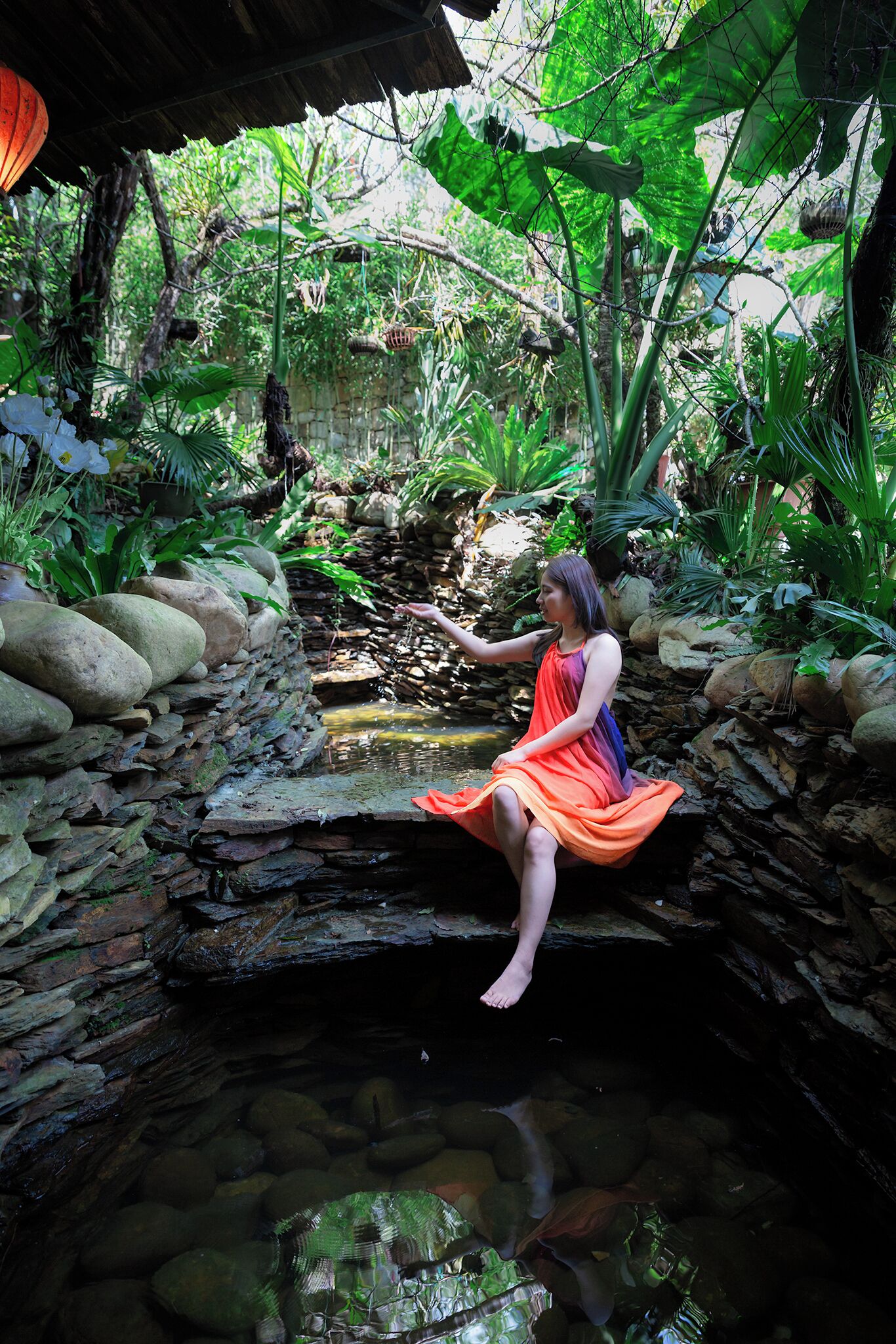 Mulher sentada em uma ponte de pedra sobre um pequeno riacho, em um jardim exuberante com pedras e plantas.