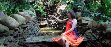 A woman sitting on a stone bridge over a small stream in a lush garden with rocks and plants.