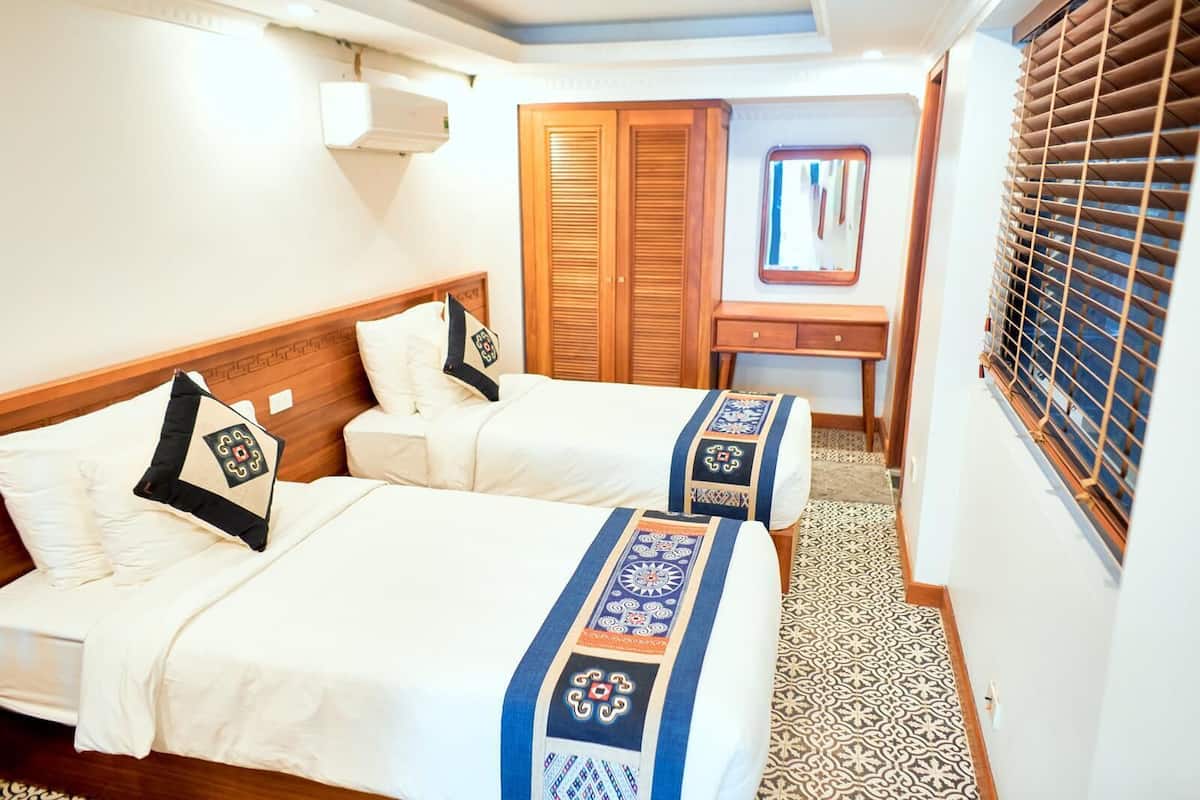 A hotel room with two beds, a wooden wardrobe, a desk, and a window with blinds.