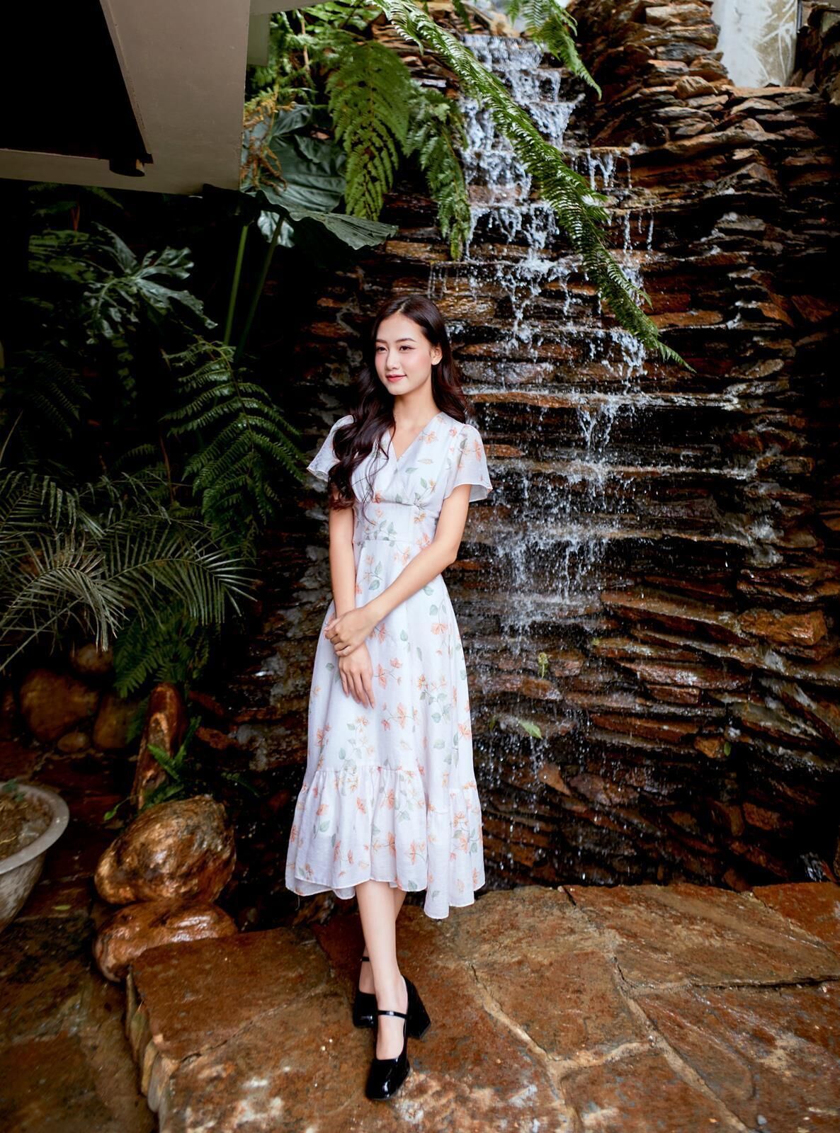 A woman standing in front of a waterfall with a rocky wall and lush greenery.