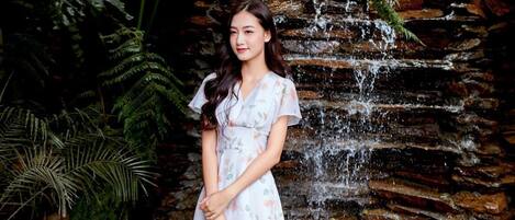 A woman standing in front of a waterfall with a rocky wall and lush greenery.