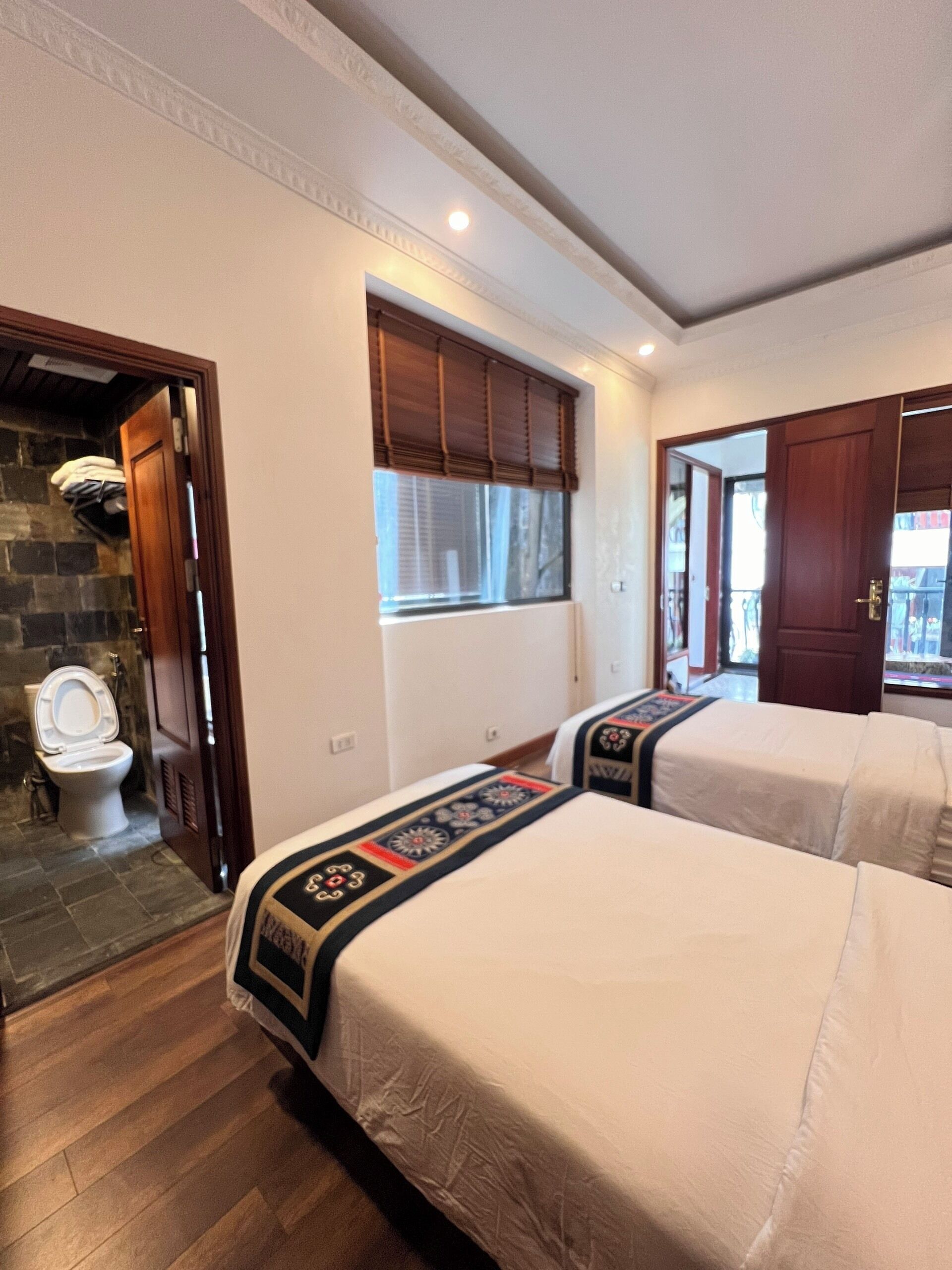 A hotel room with two beds, a bathroom with a toilet, and a window with blinds.