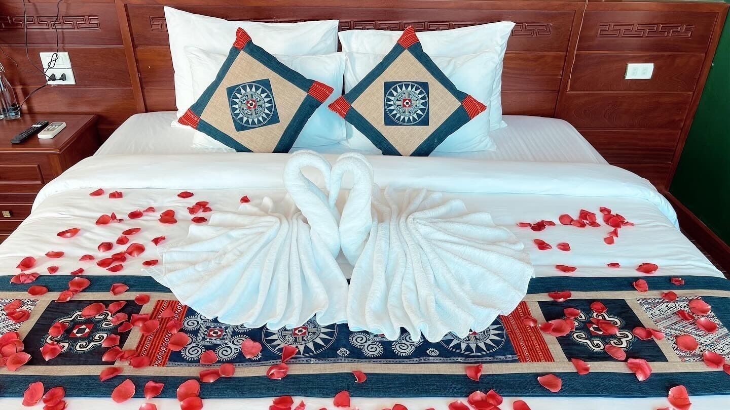 A neatly made bed with a decorative towel arrangement and rose petals, a wooden headboard, and a bedside table with a remote control.