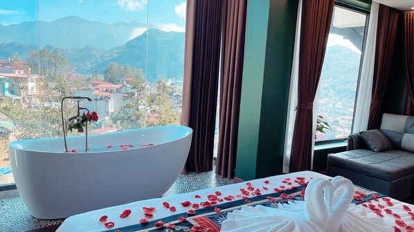 Habitación de hotel con una bañera grande llena de pétalos de rosa, una cama con pétalos de rosa y vistas a un paisaje montañoso.