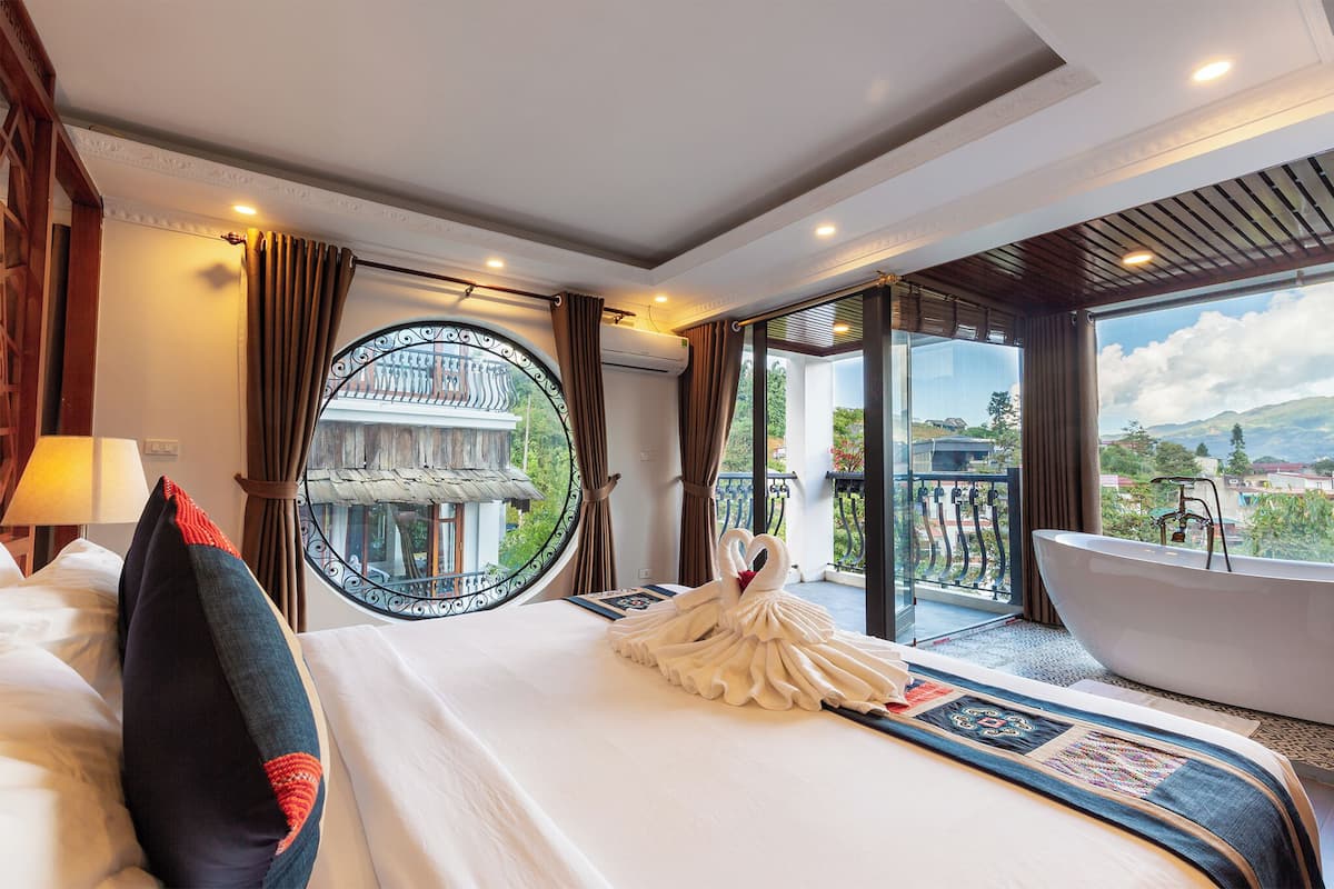 A hotel room with a large bed, a round mirror, a bathtub, and a balcony with a view of the mountains.