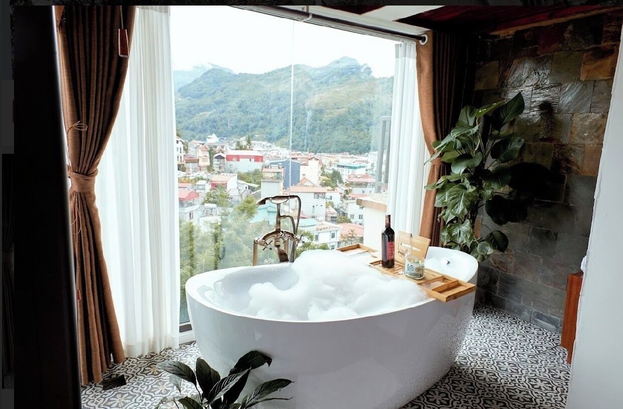 A bathroom with a large bathtub filled with bubbles, a window with a view of a cityscape and mountains, and a potted plant.