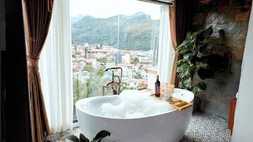 A bathroom with a large bathtub filled with bubbles, a window with a view of a cityscape and mountains, and a potted plant.