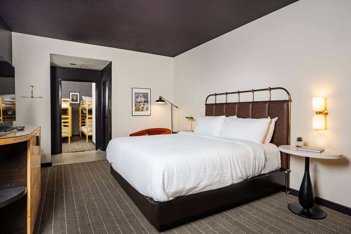 A hotel room with a large bed, a desk, a chair, a small table, and a wall-mounted lamp.