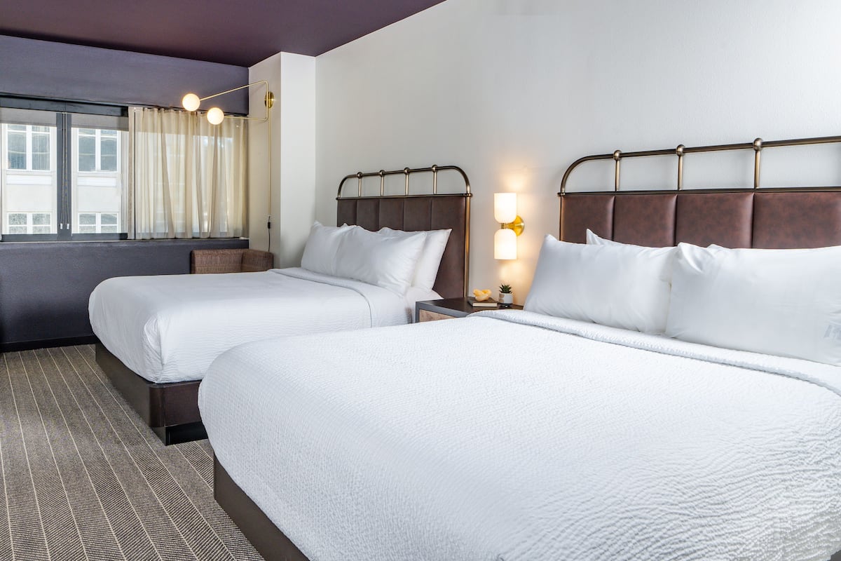 A hotel room with two beds, a window with curtains, a nightstand with a lamp, and a patterned carpet.