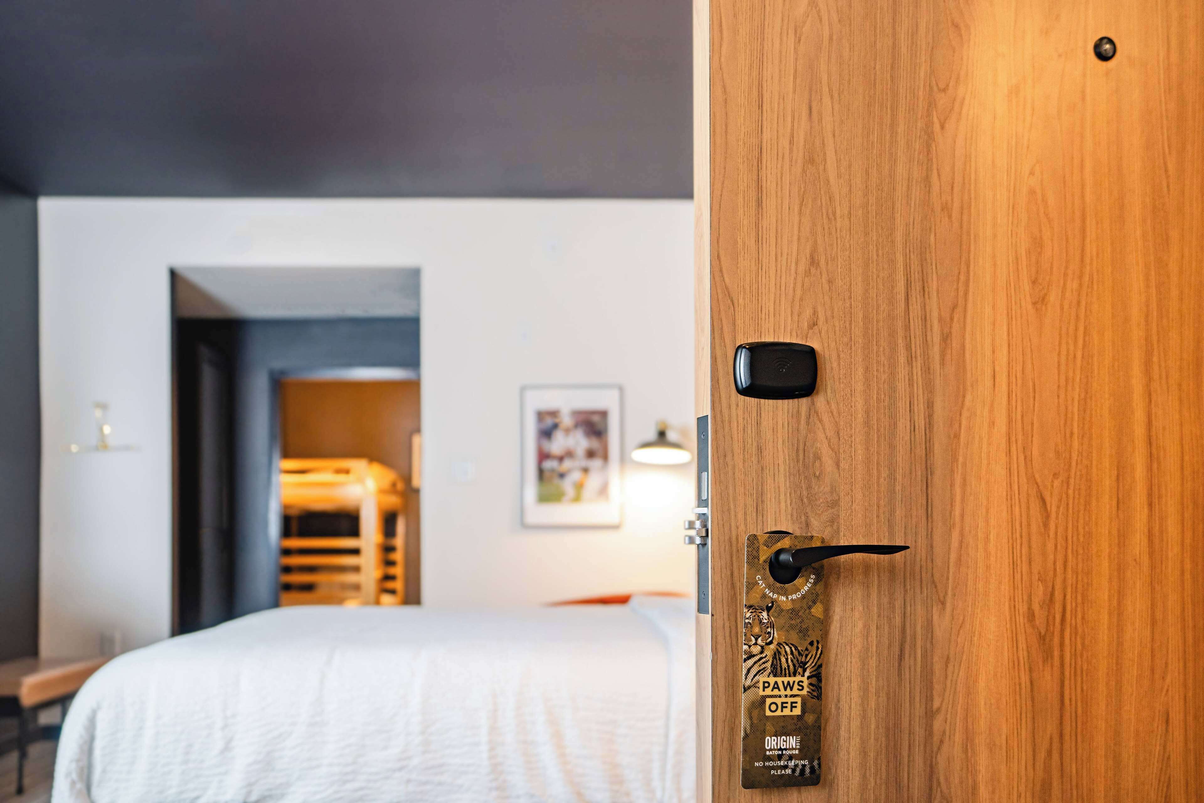 Une chambre d’hôtel avec une porte en bois, un lit recouvert d’une couette blanche, un tableau encadré au mur et une petite table avec une lampe.
