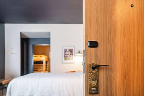 A hotel room with a wooden door, a bed with a white duvet, a framed picture on the wall, and a small table with a lamp.