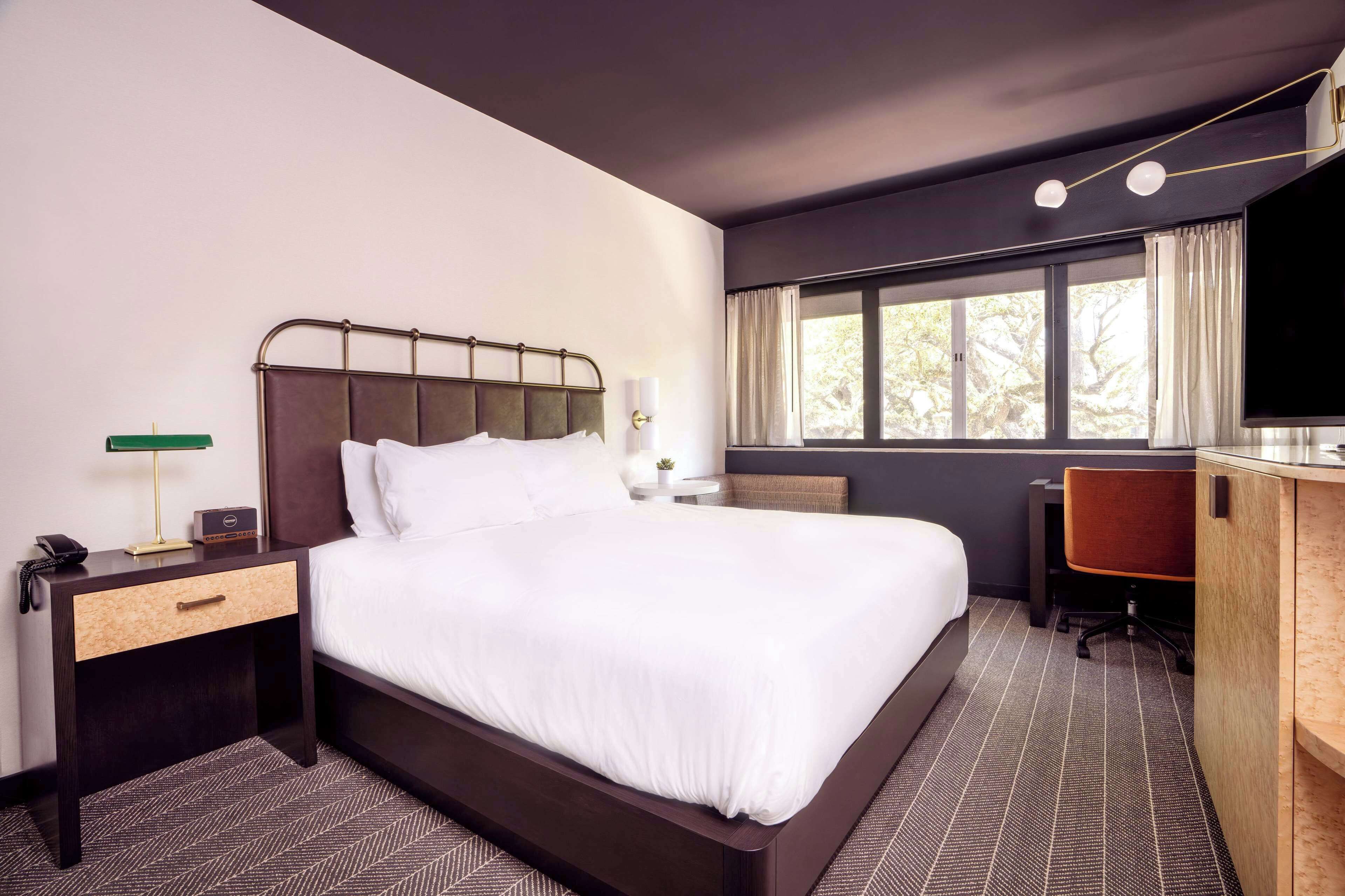 A hotel room with a large bed, a desk, a chair, a TV, and a window with curtains.