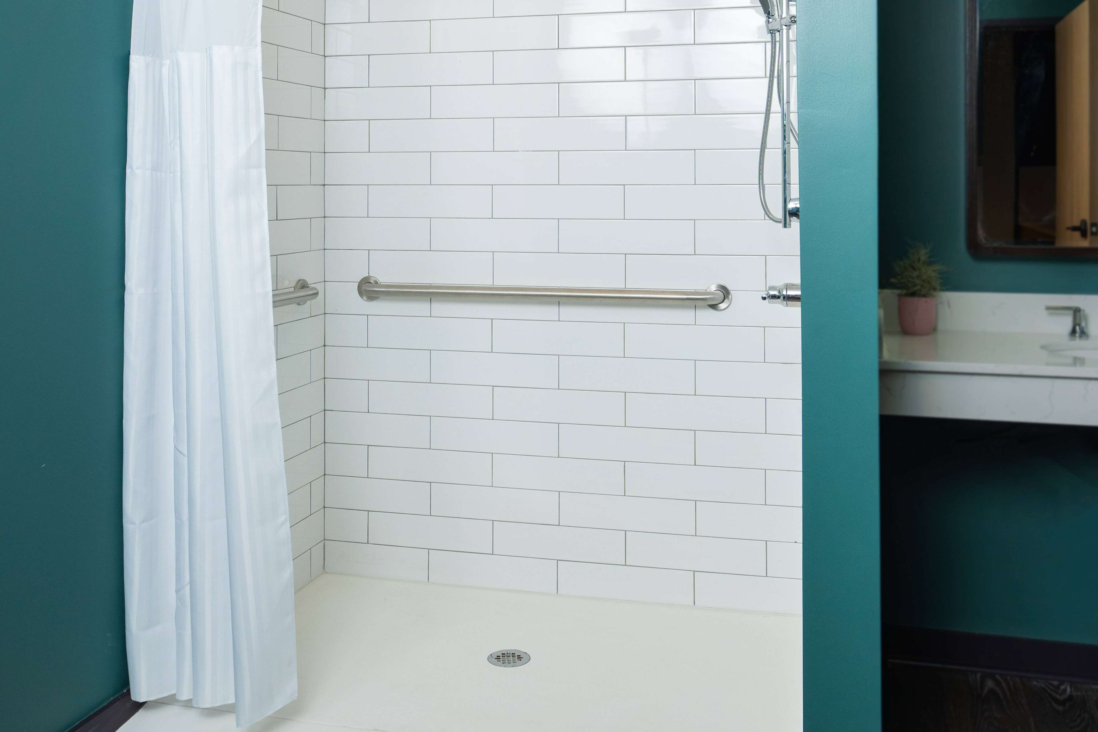A bathroom with white tiled walls, a white shower curtain, a grab bar, a sink with a mirror, and a potted plant.
